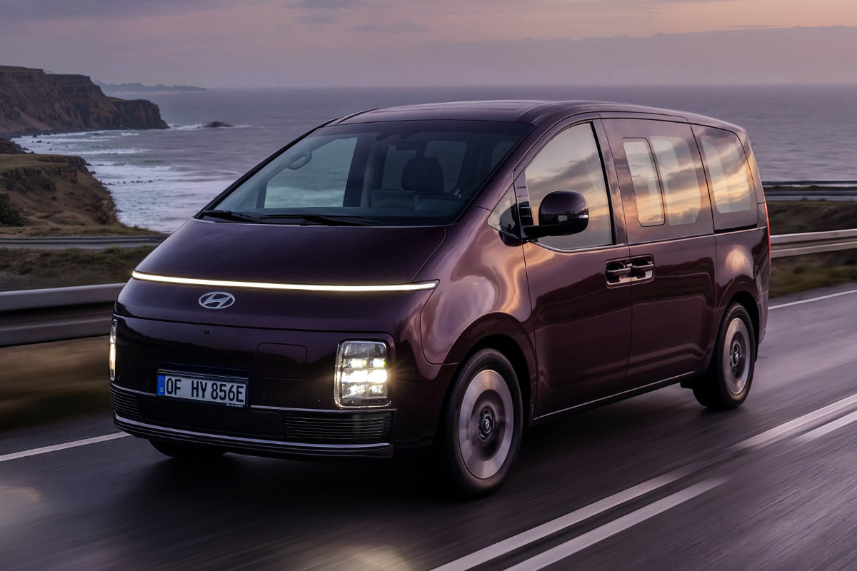 Hyundai STARIA fährt bei Sonnenuntergang auf einer Küstenstraße entlang des Meeres.